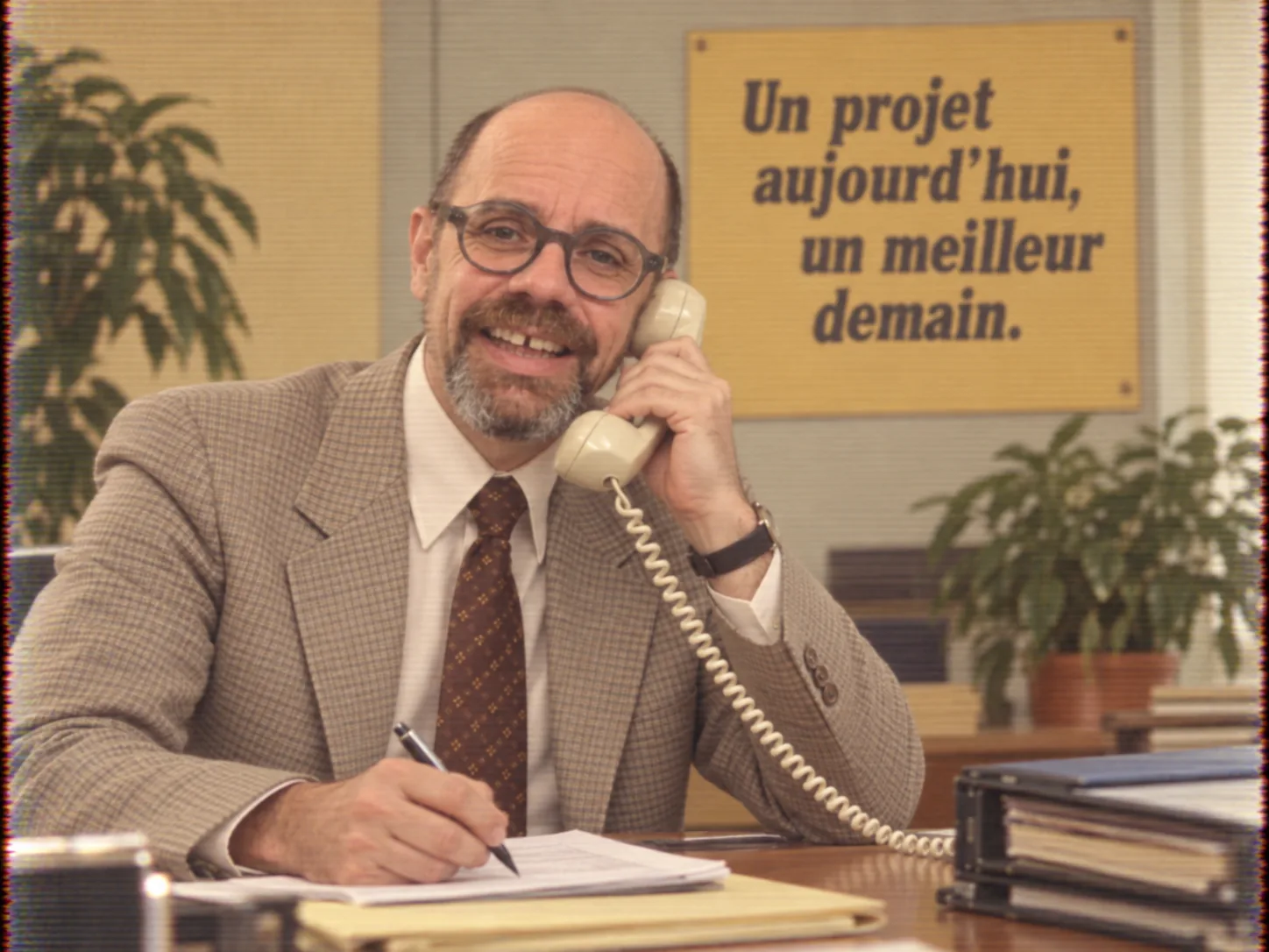 Un homme au téléphone à son bureau, sous une affiche « Un projet aujourd’hui, un meilleur demain »